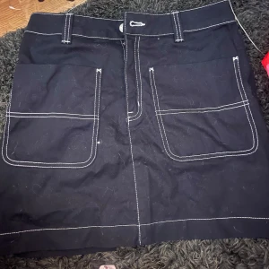 Svart kortkjol med kontrastsömmar - Svart kortkjol med stora framfickor och vita kontrastsömmar. Klassisk design med bälteshällor och dragkedja framtill. Perfekt för en chill och stilren look.