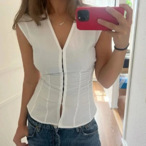Vit korsett-topp med v-ringning - Säljer en vit korsett-topp med v-ringning och markerad midja. Toppen är ärmlös och har knappar framtill som ger en snygg detalj. Perfekt att styla med jeans eller kjol för en trendig look.