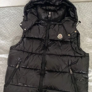 Svart dunväst från Moncler - Säljer en svart, glansig dunväst från Moncler med hög krage och avtagbar huva. Västen har två dragkedjefickor framtill, quiltad design och klassisk Moncler-logga på bröstet. Perfekt för lager-på-lager och riktigt snygg till streetwear.