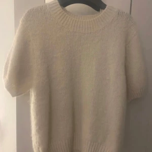 Vit stickad tshirt Gina Tricot - Mysig vit stickad tröja från Gina Tricot med korta ärmar och bred ribbad kant vid halsen. Perfekt för lager-på-lager och passar till både jeans och kjol. Enkel och stilren design som funkar året runt.