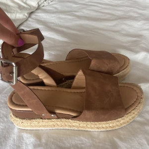 Bruna sandaler med platåsula - Snygga bruna sandaler med bred rem över foten och justerbart spänne vid ankeln. Platåsulan är klädd i flätad jute. Ovandelen är i skinnimitation och sulan är vit undertill. Perfekta för sommaren. De är dessutom supersköna att gå i! Vet ej exakt storlek eller märke då det inte står på men dem passar mig som vanligtvis har stl 39. 