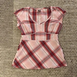 Rutig blus i rosa från TU - Supersöt rutig blus från TU i rött, rosa och vitt med v-ringning och korta ärmar. Lätt och luftig med en figurnära passform. Snygga sömdetaljer och dragkedja i sidan för enkel påklädning. Den är i bomull och skicket är jättebra förutom en väldigt liten brun prick på ryggen ungefär vid lappen som inte alls syns när man tar på sig den. Väldigt sparsamt använda och väldigt fin! Den har även lite rynkade sömmar vid armarna och på framsidan. Skriv gärna om ni har några frågor!!