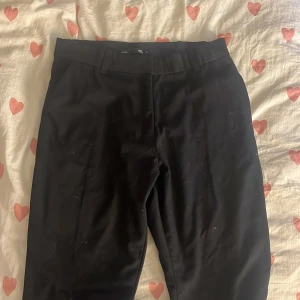 Svarta kostymbyxor från 157 - Snygga svarta kostymbyxor från 157 med klassisk rak passform och hällor för bälte. Byxorna har en stilren look och passar perfekt till en dressad outfit. Materialet känns mjukt och följsamt, vilket gör dem bekväma att bära hela dagen.