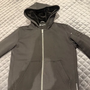 Svart hoodiejacka Moose Knuckles XL - Svart hoodiejacka från Moose Knuckles i storlek XL (BARNSTORLEK). Jackan har dragkedja framtill, två stora fickor och en ikonisk metallogo på ärmen. Insidan är fodrad med mjuk, glansig fleece för extra komfort. Perfekt för dig som gillar streetwear med premiumkänsla.