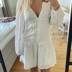 Vit broderad kortklänning med v-ringning - Superfin vit kortklänning med broderade detaljer och v-ringning. Klänningen har långa, luftiga ärmar och markerad midja med volangig kjol. Perfekt för sommarens alla tillfällen, riktigt söt och trendig stil.
