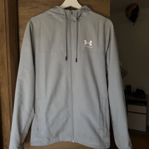 Under Armour jacka  - Säljer en stilren ljusgrå vindjacka från Under Armour med huva och dragkedja framtill. Som ny. 