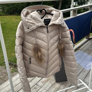 Beige dunjacka med pälskant Hollies - Snygg beige dunjacka från Hollies med huva och fluffiga pälskanter på snörena. Jackan har dragkedja framtill, två fickor med dragkedja och quiltad design. Perfekt för kyliga dagar och riktigt trendig look.