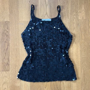Svart paljettopp från Manon - Säljer en svart glittrig topp från Manon med smala axelband och täckt av paljetter. Toppen är ärmlös och har en croppad passform som ger en snygg siluett. Perfekt för dig som vill sticka ut med lite extra bling.