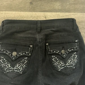 Svarta bootcut jeans med strass Madlady - Svarta low waist jeans från Madlady med bootcut-ben och snygga strassdetaljer på både fram- och bakfickor. Bakfickorna har broderade vingar med glittrande stenar. Jeansen har normal passform och är perfekta för dig som gillar att sticka ut. Säljer för de lite för stora, längden passar mej som 158, skriv för fler bilder och pris kan diskuteras 💓