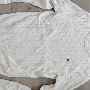 Vit kabelstickad tröja från Morris - Vit kabelstickad tröja från Morris med rund hals och diskret broderad logga på bröstet. Tröjan har klassisk kabelmönster och ribbade muddar vid ärmslut och nederkant. Perfekt för lager-på-lager och snygg till jeans eller kjol.