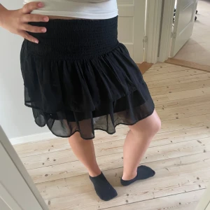 Svart volangkjol från Kappahl - Supersöt svart kortkjol från Kappahl med volanger och smock i midjan. Kjolen är luftig och har ett tunt, transparent yttertyg som ger en snygg flowy känsla. Perfekt att styla med en croppad topp eller hoodie för en chill look. Stor i storlek för mig som är vanligtvis en s.