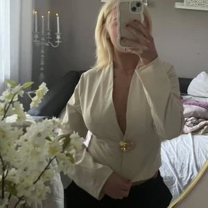 Beige kavaj topp  - Snygg beige kavaj topp med djup v-ringning och ett unikt guldigt knäppe framtill. Rak passform och långa ärmar, perfekt för dig som vill sticka ut. Stilren och enkel att matcha med både jeans och kostymbyxor.