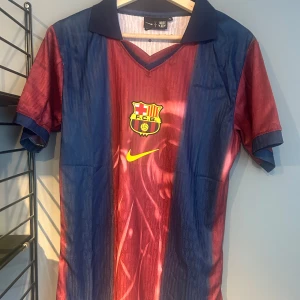 Barça x Cactus Jack fotbollströja Nike - Snygg Barcelona fotbollströja i rödblått med klassiska vertikala ränder, gul Nike-logga och FCB-emblem på bröstet. Baksidan har stort nummer 2, 'Cactus Jack' och 'BARÇA' i vitt. Skön krage och korta ärmar, perfekt för dig som vill sticka ut på planen eller gatan. Helt ny med tagg en replika