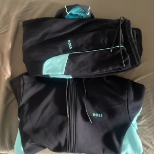 Svart och turkos Boss tracksuit - Snygg svart och turkos tracksuit från Boss med dragkedja på tröjan och matchande byxor. Setet har kontrastfärgade detaljer och Boss-logga på både överdel och byxa. Perfekt för en chill och sportig look.
