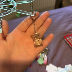 Duffy The Disney Bear nyckelring - Söt nyckelring med Duffy The Disney Bear-motiv. Nyckelringen har en brun rektangulär berlock med en glad björn på framsidan och texten 'Duffy The Disney Bear'. Perfekt att hänga på väskan eller nycklarna för en gullig touch.