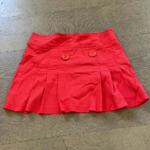 Röd lågmidjad kortkjol❤️ - Snygg röd kortkjol med två stora knappar framtill och veckad nederkant. Kjolen har dragkedja i sidan och är gjord i ett lätt bomullstyg. Perfekt för dig som vill ha en färgklick i garderoben! Så populära just nu❤️ Tryck ej köp nu