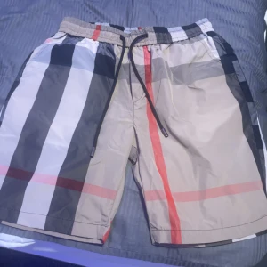 Burberry rutiga shorts med snörning - Snygga Burberry shorts med klassiskt rutigt mönster i beige, svart, vitt och röda detaljer. De har elastisk midja med svart snörning och är tillverkade i ett lätt syntetmaterial, perfekta för sommaren. Stilen är loose och de har en bakficka för extra funktion.