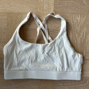 Vit linnetopp med korsade band - Snygg vit linnetopp med smala korsade axelband i ryggen. Perfekt för träning eller som ett basplagg. Mjuk och stretchig känsla med bred resår nedtill för extra stöd. Enkel och stilren design som passar till mycket.