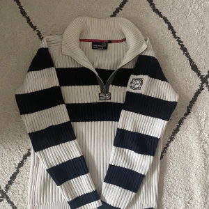 Randig stickad tröja från Pelle P - Säljer en ribbstickad tröja från Pelle P i storlek L. Tröjan är vit och marinblå med breda ränder, halv dragkedja och hög krage. Broderad logga på bröstet. Perfekt för dig som gillar sportig och klassisk stil.