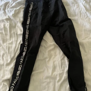 Svarta Kenzo byxor med logga - Svarta byxor från Kenzo med vit logga längs båda sidorna. De har elastisk midja med snörning och raka ben. Materialet är lätt och känns sportigt, perfekt för en chill och trendig look. Snygga detaljer med Kenzo Paris-tryck längs benen.