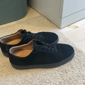 Svarta sneakers från Christian Louboutin/byte funkar  - Stilrena svarta sneakers i mocka från Christian Louboutin med klassiska röda sulor och snörning. Skorna har rund tå och platt sula, perfekt för dig som vill ha en clean look med lyxig touch. Insidan är ljus och mjuk för extra komfort.