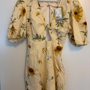 Blommig kortklänning från H&M - Superfin ljus kortklänning från H&M med blommigt mönster i gult och grönt. Klänningen har puffärmar, knytning fram och en liten cut-out vid bysten, samt öppen i ryggen. Perfekt för sommarens alla tillfällen! Tillverkad i bomull för en skön känsla.