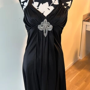 Svart långklänning från Venus med strass - Svart långklänning från Venus i glansig polyester med halterneck och snygg knytning i nacken. Framtill sitter en stor dekoration med strass och pärlor. Klänningen har draperad effekt och markerad byst. Köpt på selpy men den passa inte :/