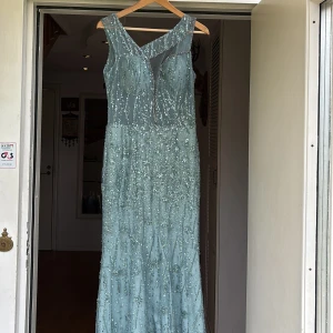 Ljusblå glittrig långklänning - Magisk ljusgrön långklänning med paljetter. Klänningen har ärmlös design, v-ringning och transparent mesh upptill. Perfekt för bal eller festliga tillfällen när du vill sticka ut.