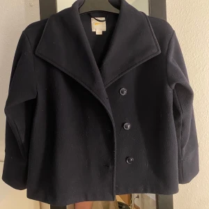 Marinblå skepparkavaj från Gina Tricot - Stilren marinblå skepparkavaj från Gina Tricot med bred krage och dubbelknäppning framtill. Jackan har en klassisk, rak passform och är tillverkad i ett mjukt ullmaterial. Perfekt för dig som vill ha en tidlös look med snygga detaljer.