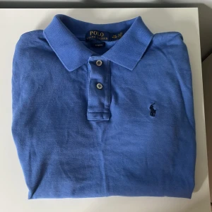 Blå pikétröja från Ralph Lauren - Snygg blå pikétröja från Polo Ralph Lauren i slim fit-modell. Den är i storlek L i barnstorlek så den passar som S i vanlig strlk. Perfekt nu till sommaren om du vill se riktigt stilren ut! Den är i väldigt fint skick, har inga skador eller fläckar.