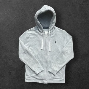 Grå hoodie från Polo Ralph Lauren - Inga hål eller fläckar! Snygg grå hoodie från Polo Ralph Lauren med dragkedja, huva med snörning och klassisk broderad logga på bröstet. Tillverkad i mjuk bomullsmix, perfekt för chill dagar. Fickor framtill och ribbade muddar för extra komfort. 