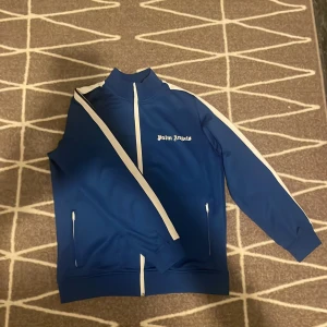 Blå vindjacka från Palm Angels - Snygg blå vindjacka från Palm Angels med vita detaljer längs dragkedja och ärmar. Jackan har hög krage, två fickor med dragkedja och klassisk logga på bröstet. Perfekt för en sportig och trendig look.