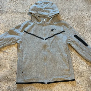 Grå Nike hoodiejacka med dragkedja XL - Snygg grå hoodiejacka från Nike i storlek XL. Jackan har hel dragkedja, huva och svarta detaljer längs bröstet och ärmen. Tillverkad i mjukt bomullsmaterial, perfekt för chill eller träning. Klassisk Nike-logga på bröstet och praktiska fickor framtill.