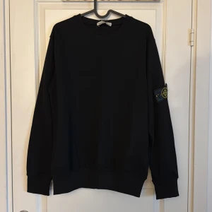 Svart Stone Island sweatshirt - Svart sweatshirt från Stone Island  med klassisk rund hals och ribbade muddar. Tröjan har den ikoniska loggan på vänster ärm och en clean, stilren look. Perfekt för dig som gillar streetwear och vill ha något enkelt men ändå exklusivt.