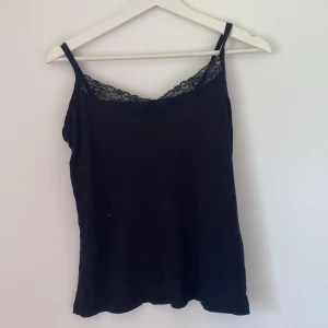 Svart linnetopp med spetskant - Svart linnetopp med tunna axelband och snygg spetsdetalj längs v-ringningen. Enkel och stilren modell som passar perfekt till jeans eller kjol. Mjuk och lätt i materialet, perfekt för varma dagar eller lager på lager.