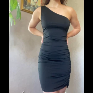 Svart one shoulder kortklänning - Superfin svart one shoulder-klänning med tight passform. Klänningen har rynkade detaljer längs sidorna, vilket ger en väldigt smickrande passform! Materialet är följsamt och lite ”glansigt”. Använd ett fåtal gånger. 
