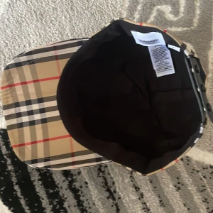 Burberry rutig keps i bomull - Snygg keps från Burberry med klassiskt beige, svart, vit och röd rutigt mönster. Justerbar baktill med svart spänne och tydlig Burberry London England-patch. Tillverkad i bomull och polyester, perfekt för att lyfta din streetstyle.