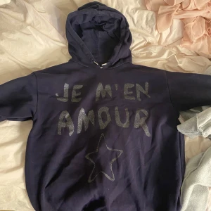 Mörkblå hoodie med glittrig text - Säljer en mörkblå hoodie med huva och glittrig texten 'JE M'EN AMOUR' på framsidan samt en stjärna under. Tröjan har en avslappnad passform och är perfekt för dig som gillar statement-plagg. Supermjuk och skön att ha på sig.