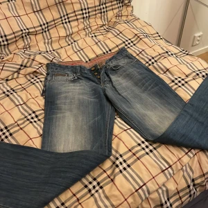 Dolce & Gabbana blå jeans med detaljer - Snygga blå jeans från Dolce & Gabbana med slitna detaljer och coola kontrastsömmar på bakfickorna. Klassisk femficksmodell med knappar framtill och unika badges samt etikett bak. Perfekta för dig som vill sticka ut med designerjeans! Jag har inte använt dom här en enda gång och dom är i ny skick! Vi kan diskutera om priset!