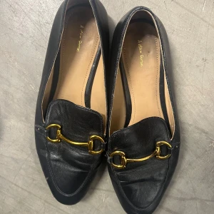 Svarta loafers med guldspänne - Stilrena svarta loafers i skinn från & Other Stories med stilrent guldfärgat metallspänne. Skorna har platt sula och rundad tå, perfekt för en klassisk och dressad look. 