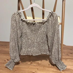 Leopardmönstrad blus - Supersöt blus från Sisters Point med leopardmönster i beige och brunt. Smockad överdel, långa puffiga ärmar med breda smockade muddar och volangdetalj vid halsringningen. Perfekt för dig som vill sticka ut lite extra.
