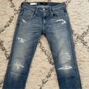 Replay Anbass blå jeans med slitningar - Eftertraktade snygga blå jeans från Replay, modell Anbass, med coola slitningar och tvättad look. Klassisk femficksdesign, normal passform och mellanblå färg. Jeansen har läderpatch med Replay-logga bak och detaljerade sömmar på bakfickan. Hör av er vid minsta lilla!! 🙌
