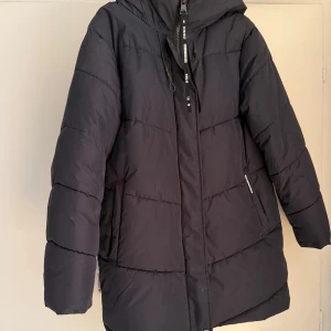 Svart pufferjacka från Khujo - Svart pufferjacka från Khujo med stor huva, dragkedja och snörning. Jackan är quiltad och har en clean, oversized look med diskreta detaljer och praktiska fickor. Perfekt för kalla dagar när du vill hålla stilen och värmen.