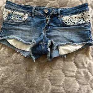 Superdry jeansshorts med spetsdetaljer - Super snygga blå jeansshorts från Superdry med råa kanter och vita virkade spetsdetaljer vid fickorna samt där bak. Shortesen är lågmidjade och har en klassisk femficksdesign. I storlek XS🩷