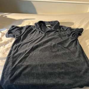 Mörkblå frotté pikétröja Kappahl - Mjuk och chill mörkgrå pikétröja från Kappahl i frotté. Klassisk krage och korta ärmar, perfekt för en avslappnad stil. Skön att bära under varmare dagar eller efter träning. Enkel och stilren design som funkar till det mesta.