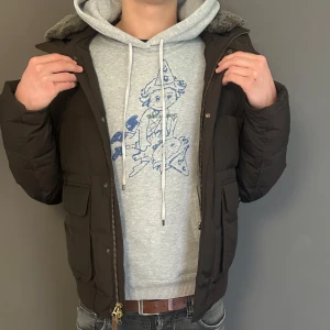 Brun pufferjacka från Woolrich - Säljer en brun pufferjacka från Woolrich med ribbade muddar, hög krage med grå fuskpäls och stora fickor framtill. Jackan har dragkedja och tryckknappar, samt en patch med Woolrich-logga på ärmen. Perfekt för kalla dagar.