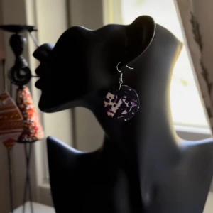 Svart och vitmönstrat örhänge från Shop with Tyra - Coolt, runt örhänge med svart och vitt mönster och inslag av lila. Hängande modell med metallkrok, perfekt för dig som vill sticka ut med ett unikt smycke. Designen är konstnärlig och edgy, passar dig som gillar statement-accessoarer. Följ oss för mer på Instagram; @shop_with_tyra