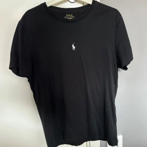 Svart t-shirt från Polo Ralph Lauren - Svart t-shirt från Polo Ralph Lauren med klassisk passform och rund hals. Ikoniska vita broderade loggan på bröstet ger en clean och stilren look. Tillverkad i mjuk bomull som känns skön mot huden. Perfekt till jeans eller shorts.