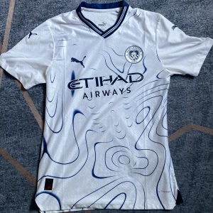 Manchester City tröja - Manchester City tröja från Puma i vitt med blå abstrakta mönster och klubbmärke på bröstet. Tröjan har korta ärmar, V-ringad krage med blå detaljer och är tillverkad i lätt, ventilerande funktionsmaterial. Etihad Airways sponsortryck på framsidan.