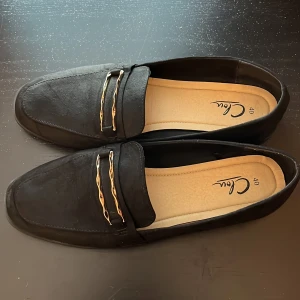 Svarta loafers med gulddetaljer från Clou - Stilrena svarta loafers från Clou i mocka-look med fyrkantig tå och två vågiga guldfärgade metalldetaljer över vristen. Skorna har platt sula och en klassisk design som passar till många outfits.